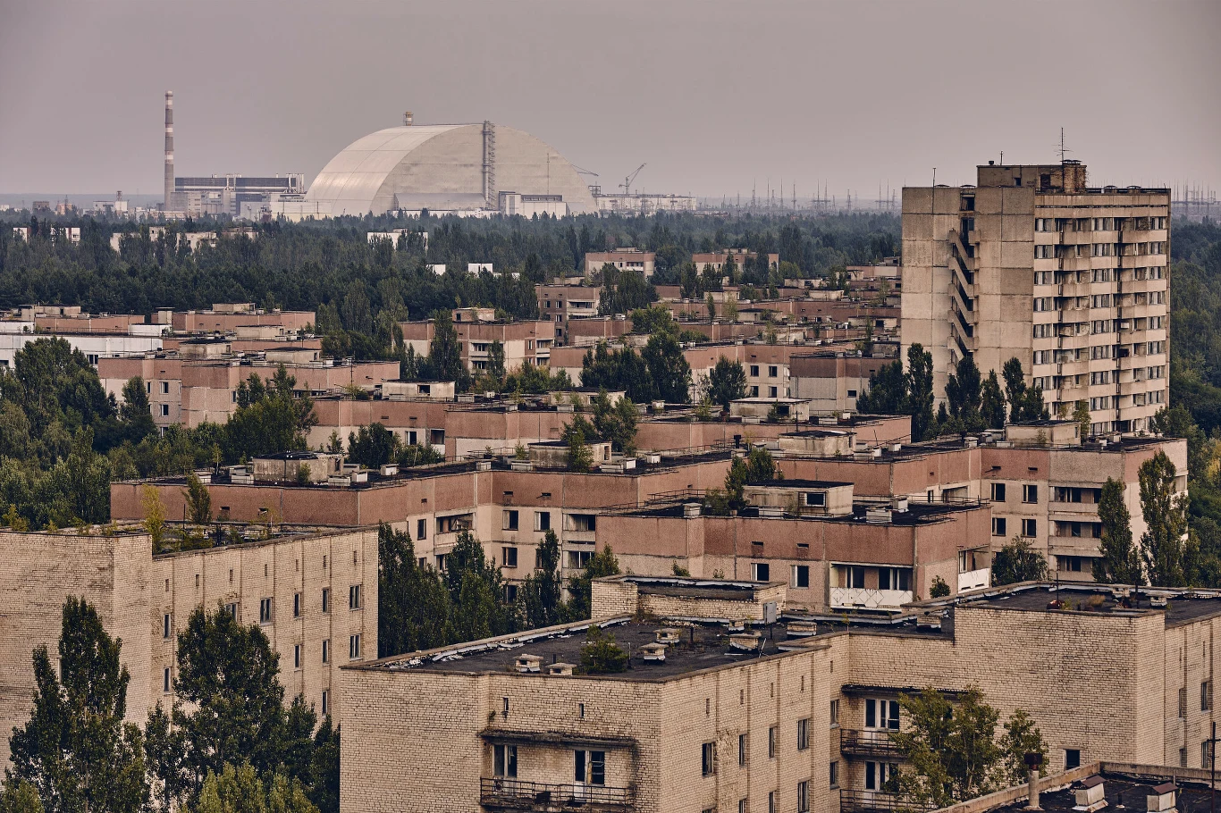 Prypeć - miasto zbudowane dla pracowników elektrowni w Czarnobylu Opuszczone blokowiska z płaskimi dachami, pokryte roślinnością, w tle charakterystyczna kopuła sarkofagu reaktora oraz komin elektrowni jądrowej otoczone gęstym lasem.