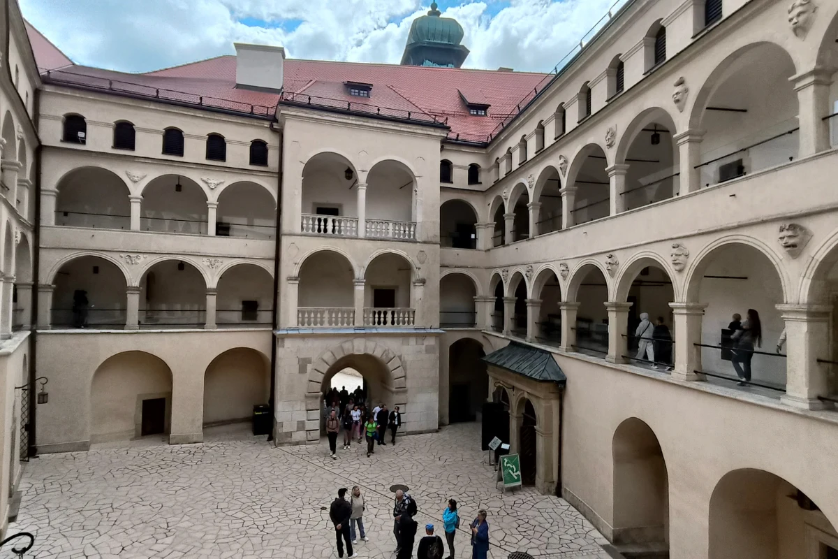 Renesansowy dziedziniec zamkowy otoczony dwupoziomowymi krużgankami z arkadami, pośrodku brukowana posadzka, w tle ludzi zwiedzających oraz wejście prowadzące do wnętrza budynku.