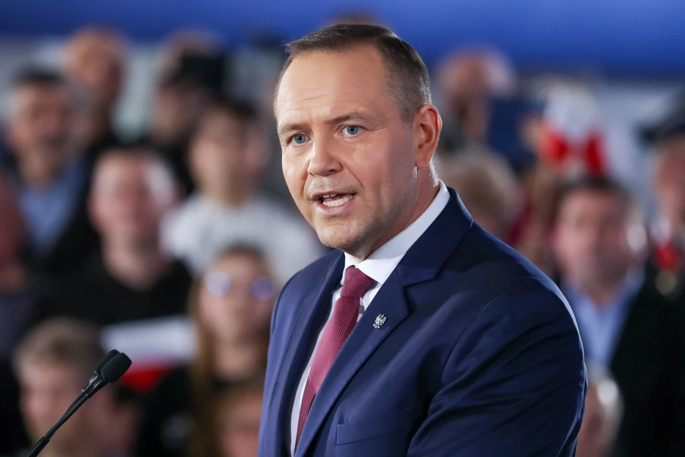 Karol Nawrocki Karol Nawrocki w garniturze i czerwonym krawacie przemawia do mikrofonu przed tłumem z biało-czerwonymi flagami