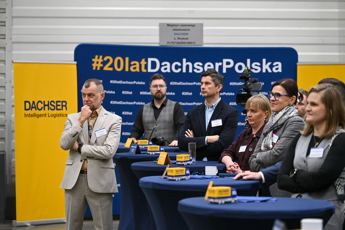 kilka osób uczestniczących w oficjalnym wydarzeniu firmowym, stojących przy wysokich stołach, na których znajdują się miniaturowe ciężarówki oraz gadżety promujące markę Dachser Polska, tło stanowi niebieska ściana z napisem #20latDachserPolska