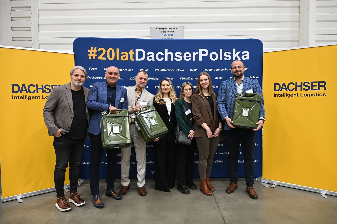siedem osób w eleganckich ubraniach pozuje na tle żółto-niebieskiej ścianki z napisem #20latDachserPolska, trzech mężczyzn trzyma duże zielone plecaki z widocznym logotypem, po bokach dominują reklamy firmy Dachser Intelligent Logistics