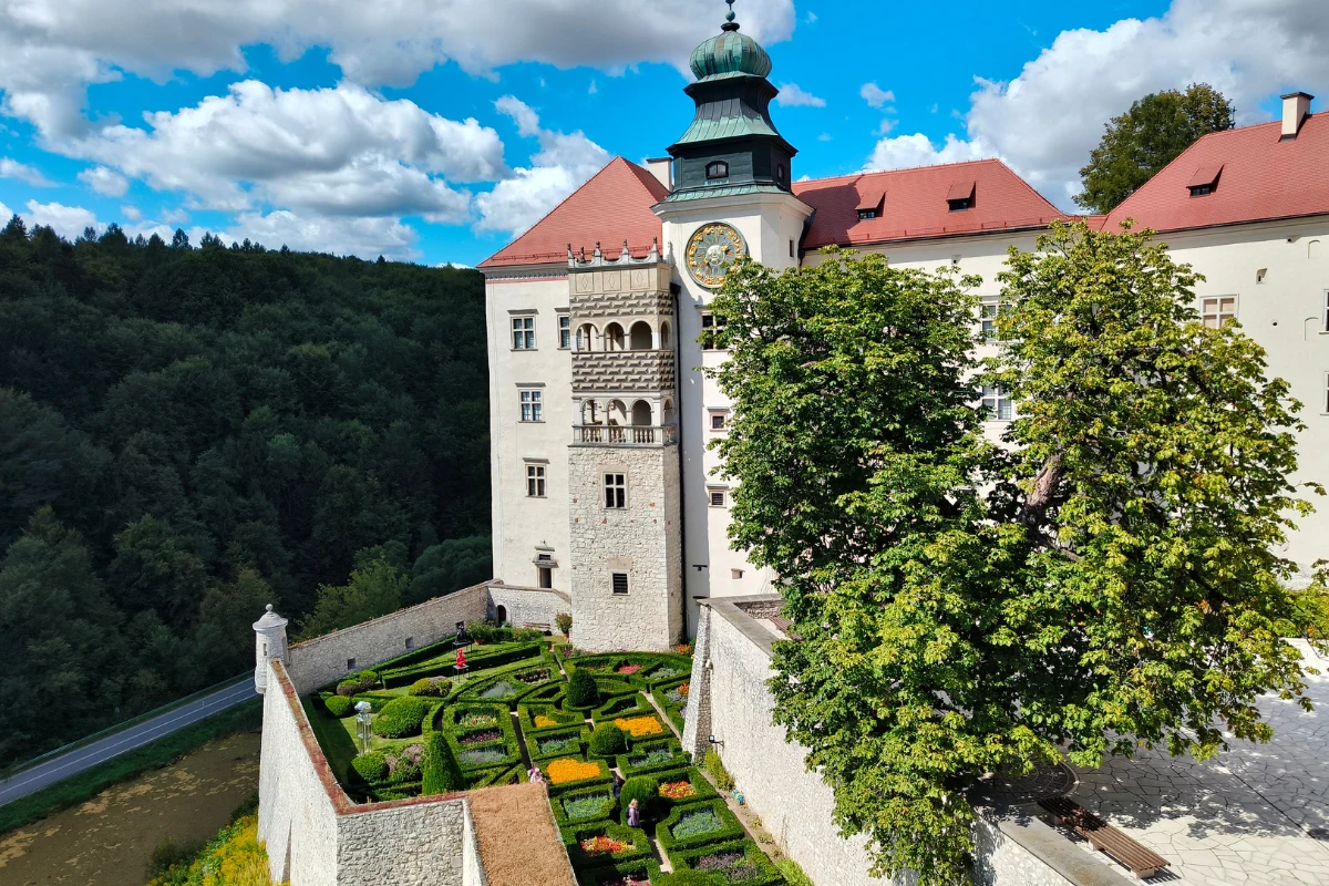 zamek z wysoką wieżą zegarową, otoczony geometrycznym ogrodem z ciekawymi rabatami i niskimi żywopłotami, w tle gęsty las oraz fragment murów obronnych