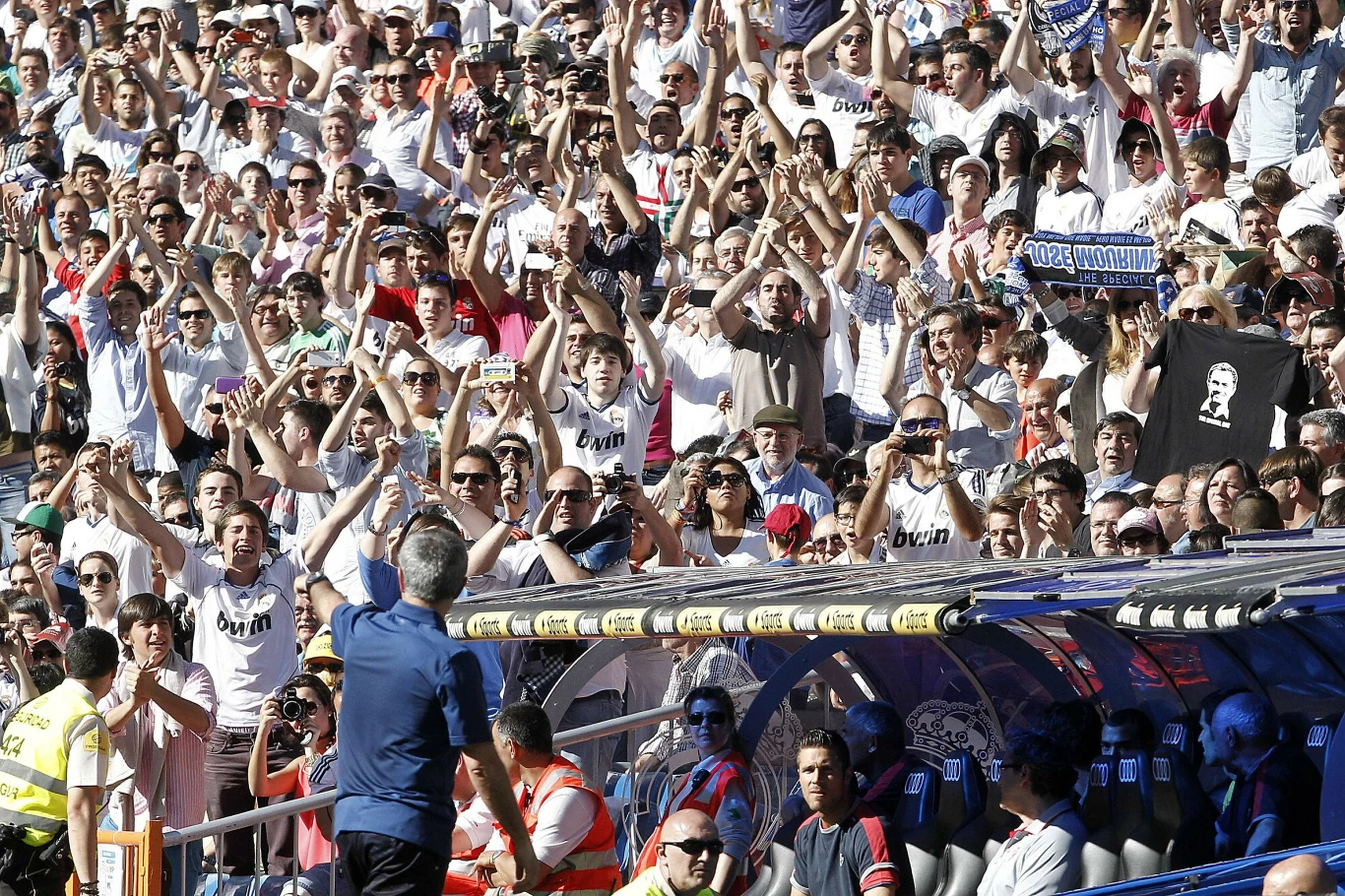 Jose Mourinho po ostatnim meczu w Realu Madryt, 01.06.2013 r. tłum kibiców ubranych głównie na biało, podnoszących ręce i wiwatujących podczas meczu piłkarskiego na stadionie, w centrum trener drużyny zbliżający się do trybun, w tle ławka rezerwowych ze sztabem szkoleniowym i piłkarzami