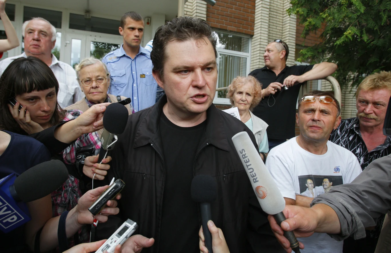 mężczyzna w ciemnej kurtce otoczony przez grupę dziennikarzy trzymających mikrofony różnych stacji telewizyjnych, w tle tłum ludzi oraz policjant