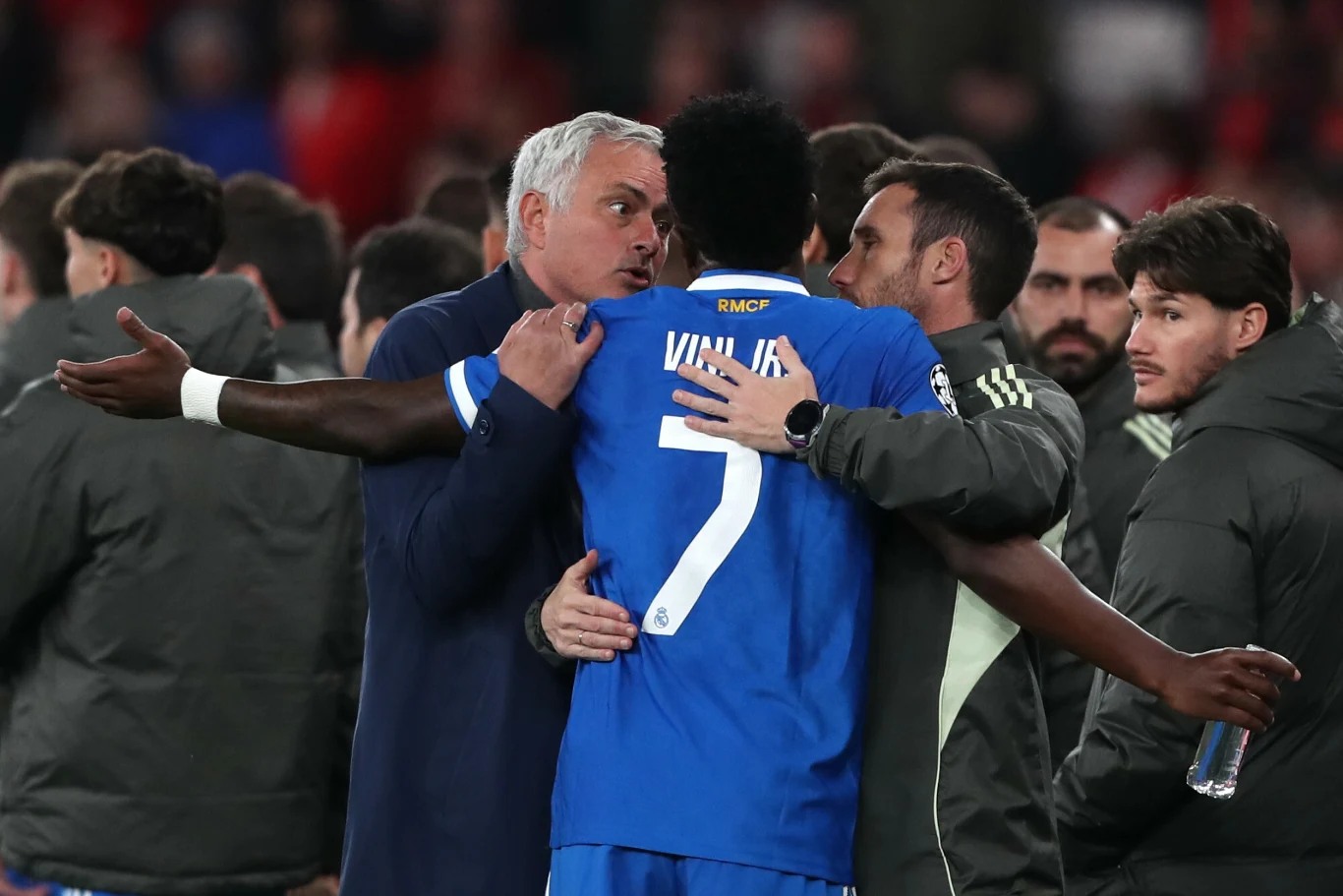 Jose Mourinho Jose Mourinho w płaszczu obejmuje piłkarza Viniciusa, w tle członkowie drużyny i rozmazany tłum kibiców.