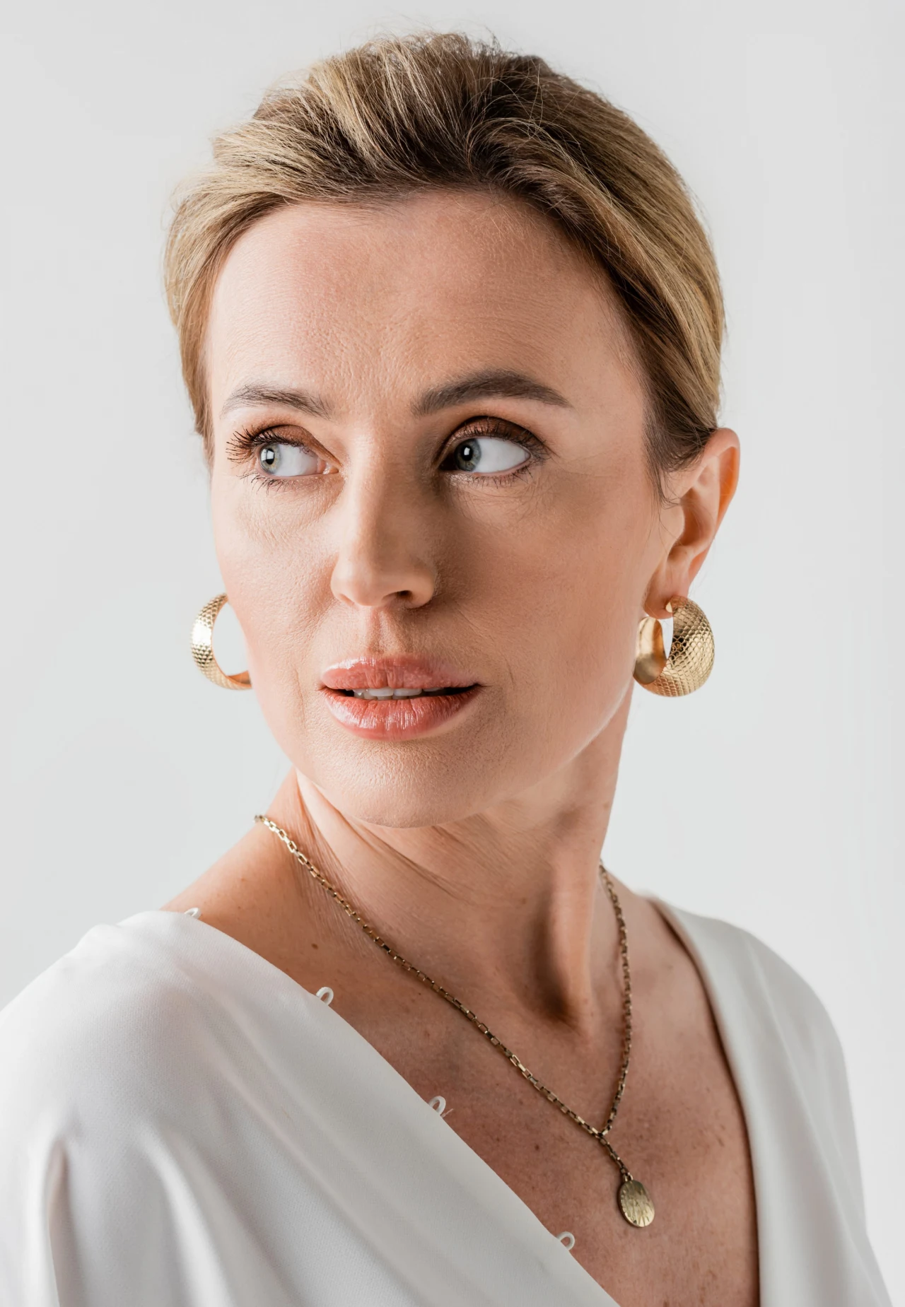 Kobieta z blond włosami upiętymi do tyłu, ubrana w białą bluzkę z dekoltem, ozdobiona dużymi złotymi kolczykami i delikatnym naszyjnikiem na szyi, spogląda w bok z poważnym wyrazem twarzy.