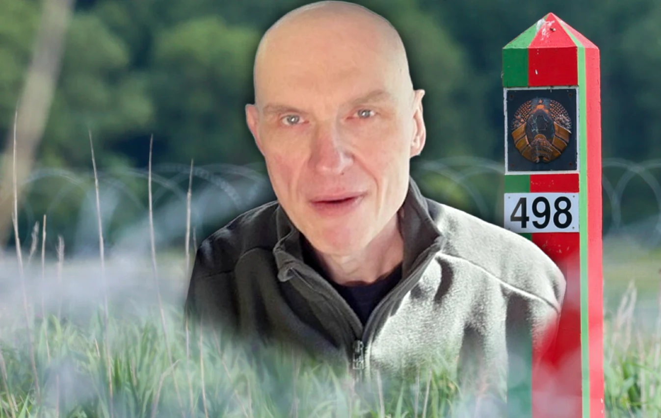 łysy mężczyzna w kurtce stoi obok słupa granicznego oznaczonego numerem 498, z widocznym orłem i kolorami zielonym, czerwonym oraz białym; w tle rozmyte ogrodzenie z drutu kolczastego i las