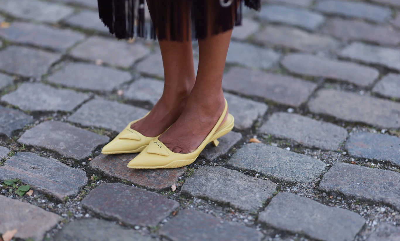 Modne buty do eleganckiej sukienki na komunię żółte czółenka na niskim obcasie na stopach osoby stojącej na brukowanej ulicy, widoczny dolny fragment ciemnej spódnicy