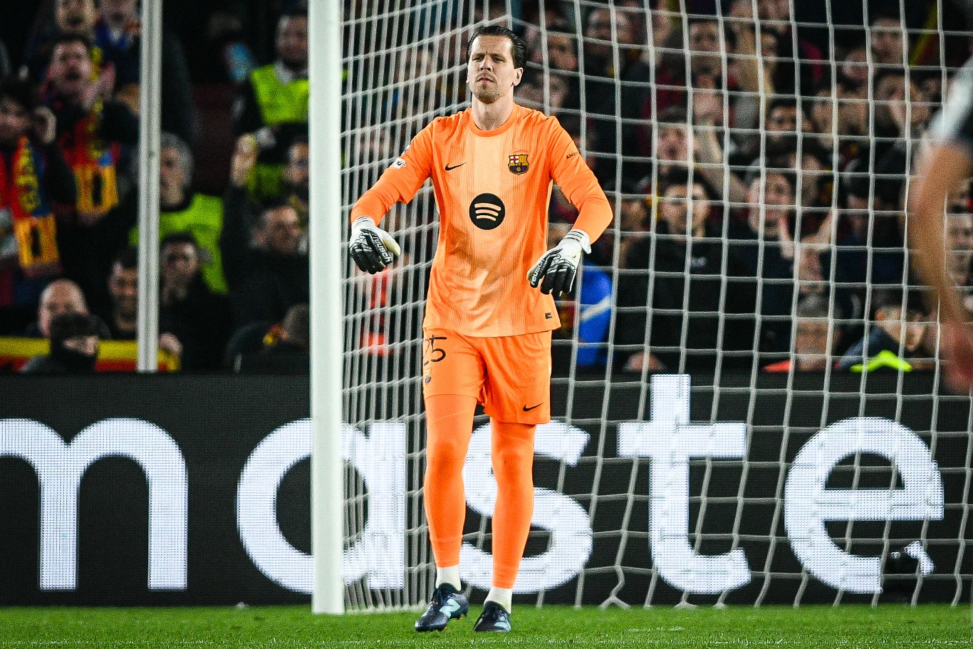 Wojciech Szczęsny bramkarz w pomarańczowym stroju z rękawicami stoi przed bramką na tle siatki i tłumu kibiców na stadionie piłkarskim