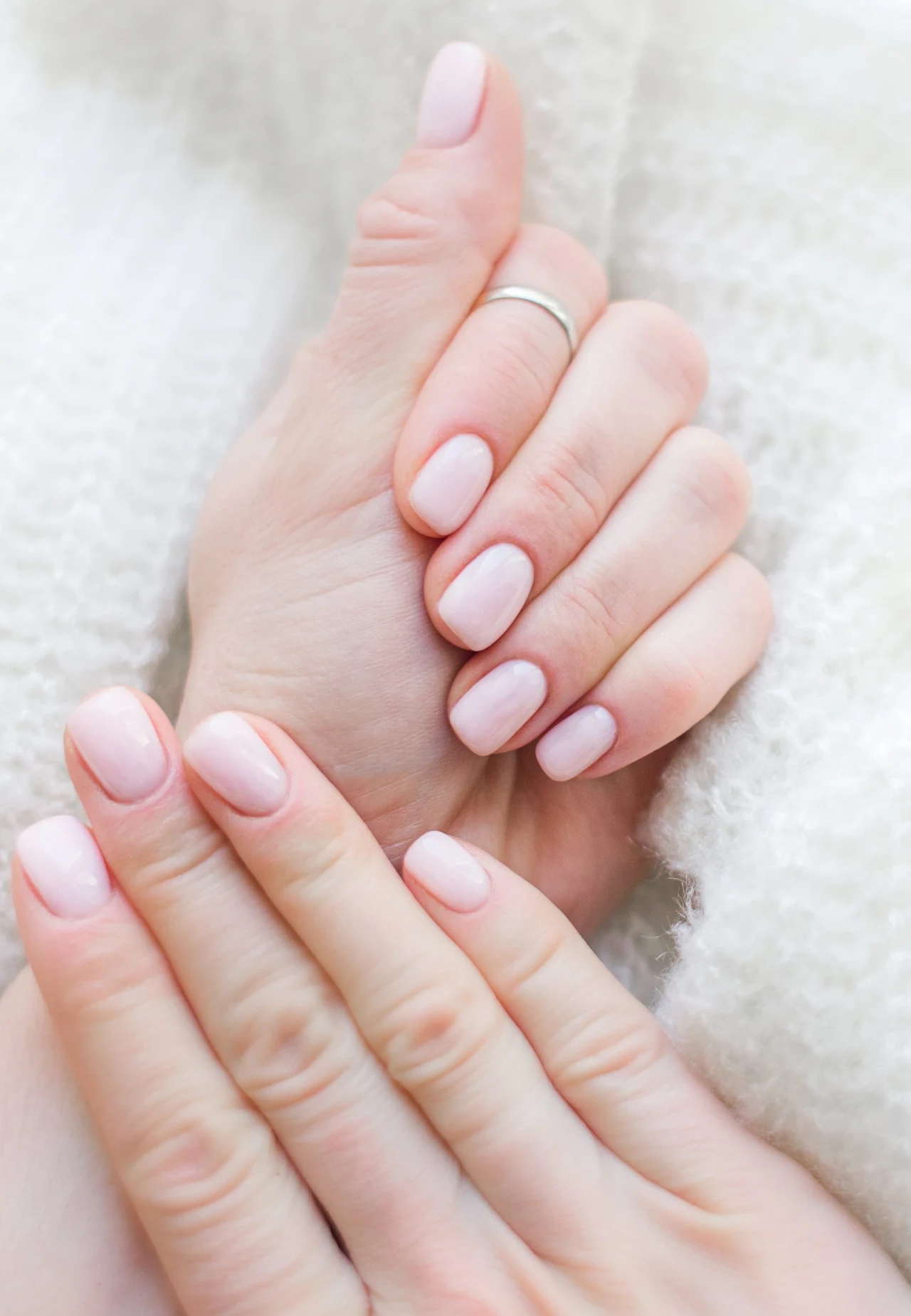 Zadbane dłonie z krótkimi, jasnoróżowymi paznokciami trzymane delikatnie na tle białego, miękkiego swetra, na jednym z palców widoczna srebrna obrączka.