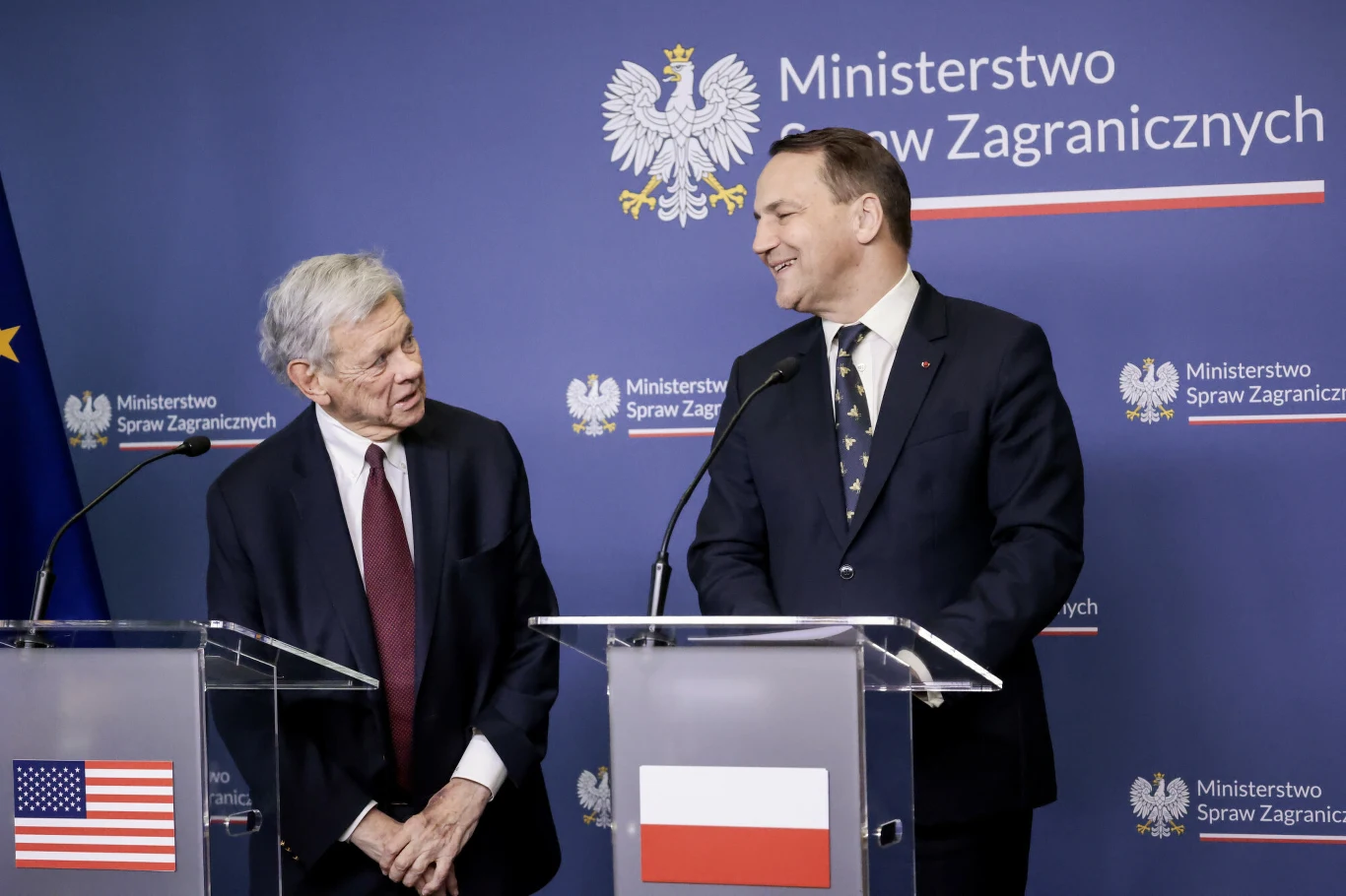dwóch mężczyzn w garniturach stoi obok siebie przy mównicach z flagą Stanów Zjednoczonych i Polski, w tle emblemat Ministerstwa Spraw Zagranicznych oraz godło Polski