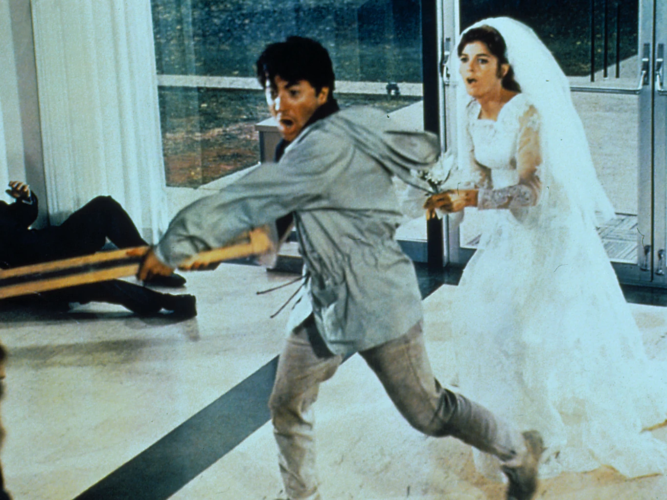 Dustin Hoffman i Katherine Ross w filmie "Absolwent" mężczyzna w jasnej kurtce biegnie przez pomieszczenie, trzymając duży drewniany kij, a za nim podąża kobieta w sukni ślubnej i welonie, wyraźnie zaniepokojona