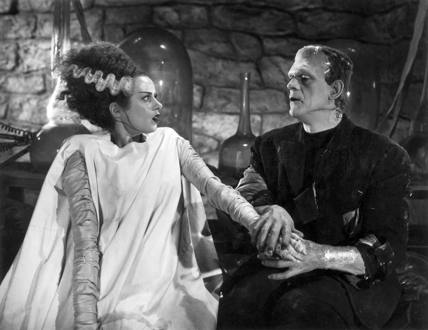 Elsa Lanchester i Boris Karloff w filmie "Narzeczona Frankensteina" kobieta z charakterystycznie ułożonymi włosami oraz mężczyzna o potężnej sylwetce i szwach na twarzy siedzą obok siebie, trzymając się za ręce, w tle kamienna ściana i laboratoryjne szkło