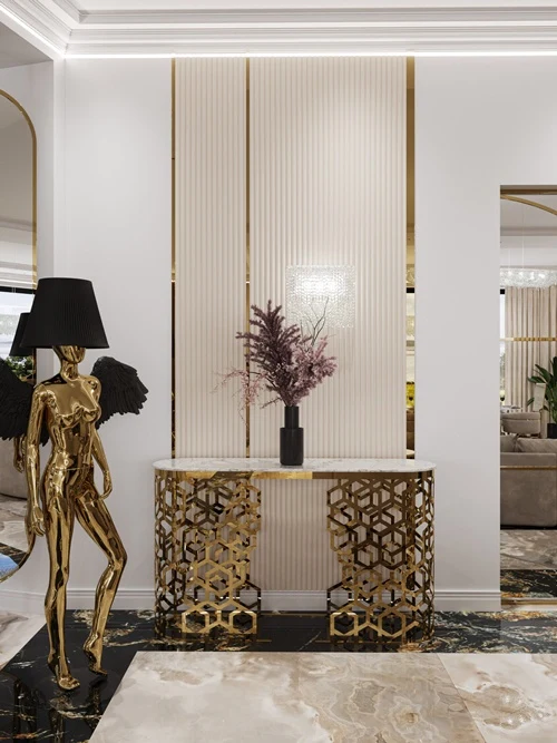 Złota rzeźba postaci z kloszem lampy zamiast głowy i czarnymi skrzydłami ustawiona obok konsoli z geometrycznym, złotym stelażem, na której stoi czarna waza z suchymi gałązkami, tło stanowi elegancka, jasna ściana z pionowymi dekoracyjnymi panelami.