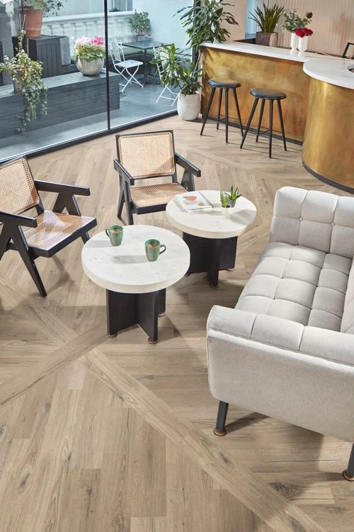 Nowoczesny salon z jasnoszarą sofą, dwoma fotelami z plecionymi oparciami, dwoma okrągłymi stolikami, na których stoją kubki i doniczka, oraz złotym barem z wysokimi krzesłami w tle; przez szklaną ścianę widoczny taras z roślinami.