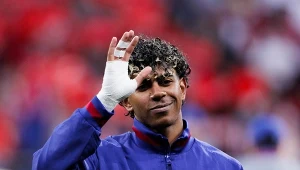 piłkarz w niebieskiej kurtce FC Barcelona wykonuje gest dłonią, na której ma opatrunek bandażowy, na tle rozmytych, czerwonych barw trybun stadionu