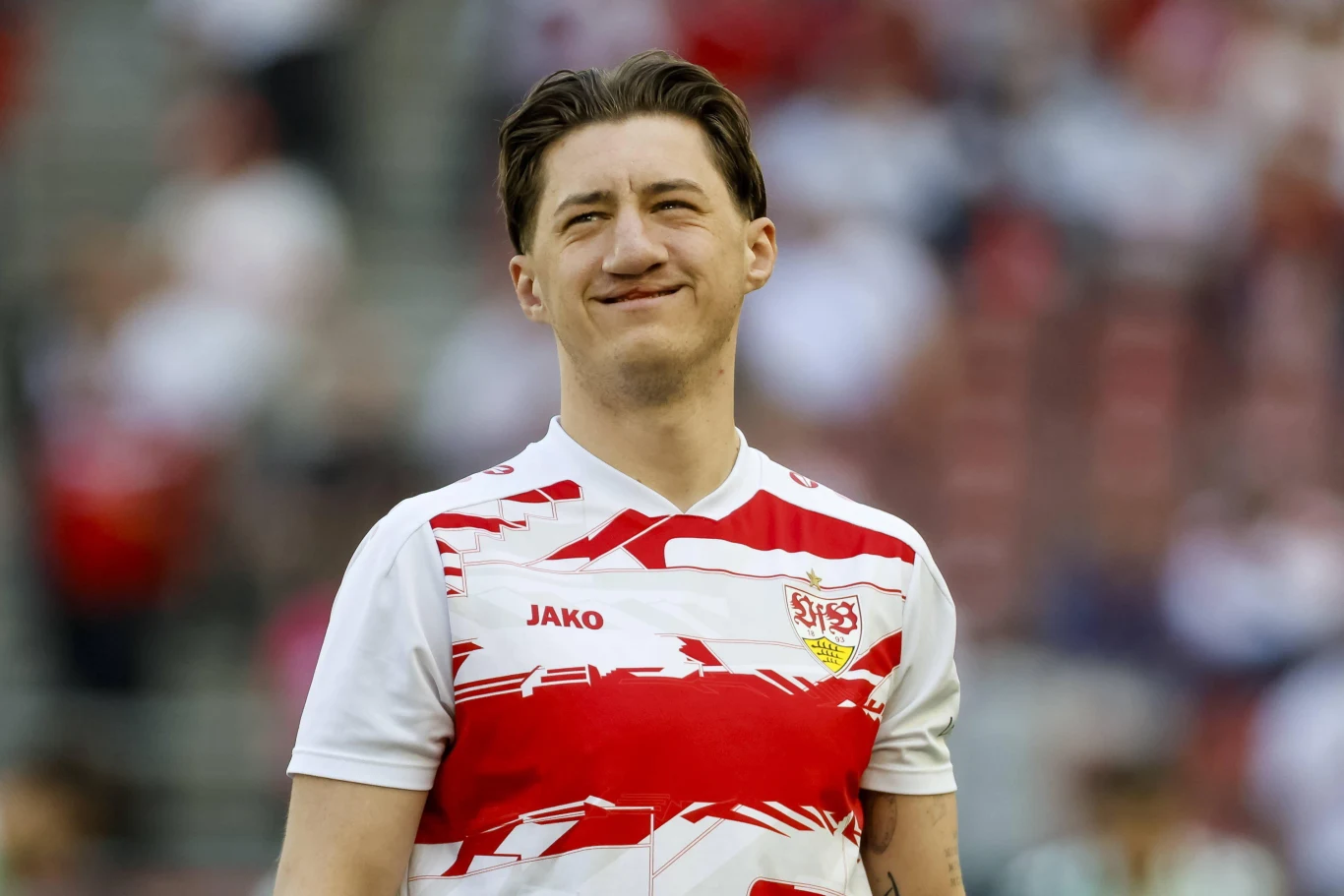 młody mężczyzna w sportowej koszulce VfB Stuttgart z widocznym logo klubu i producenta, uśmiechnięty, stoi na stadionie w otoczeniu rozmazanego tłumu