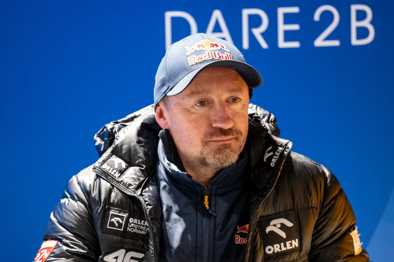 mężczyzna w ciemnej kurtce zimowej z logotypami marek Orlen, Red Bull i Dare2b, noszący czapkę z daszkiem Red Bulla, siedzi na tle niebieskiej ściany z dużym napisem DARE2B