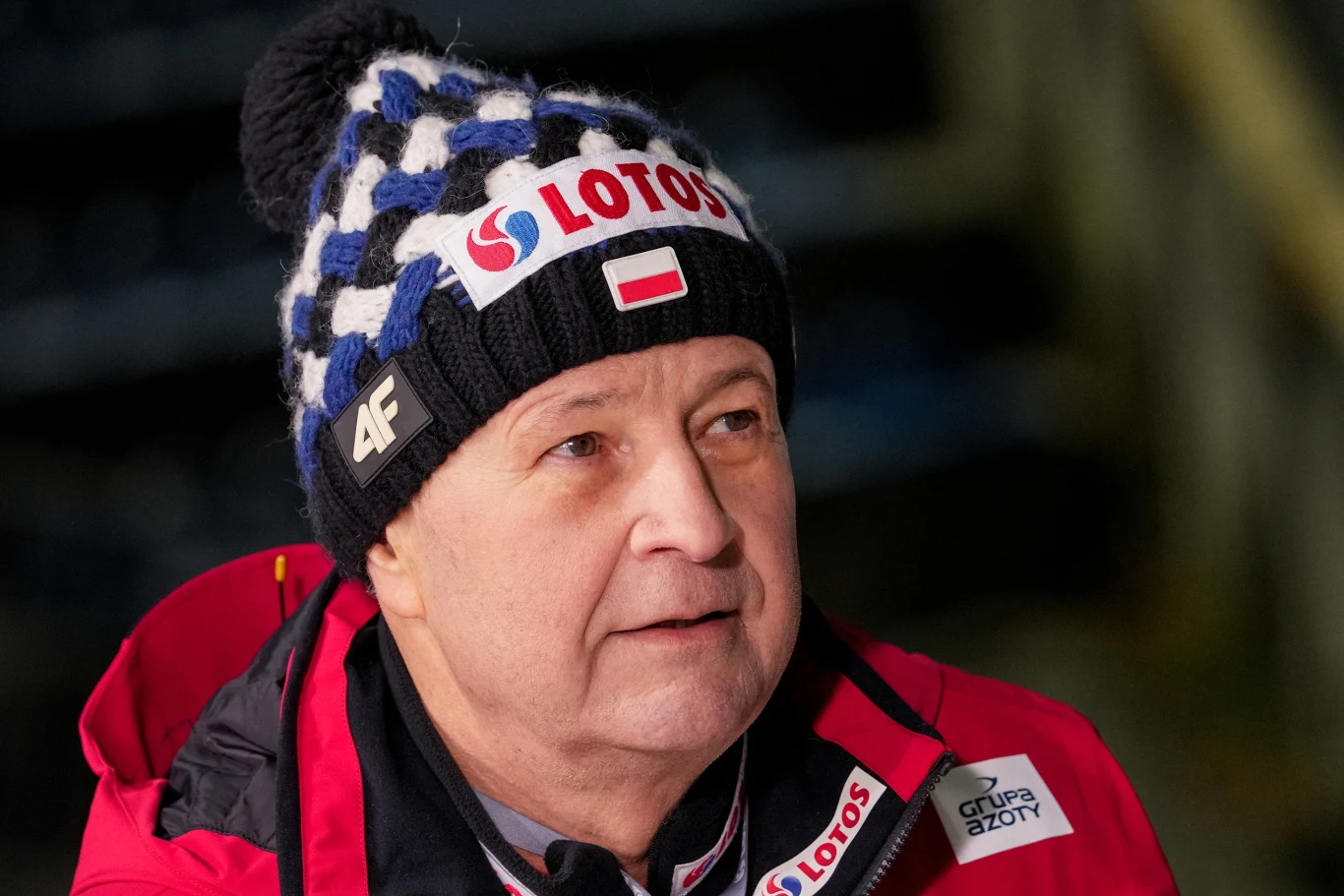 Mężczyzna w zimowej czapce z napisem 'Lotos', flagą Polski i logo 4F oraz kurtce sportowej, spogląda w bok w neutralnym otoczeniu.