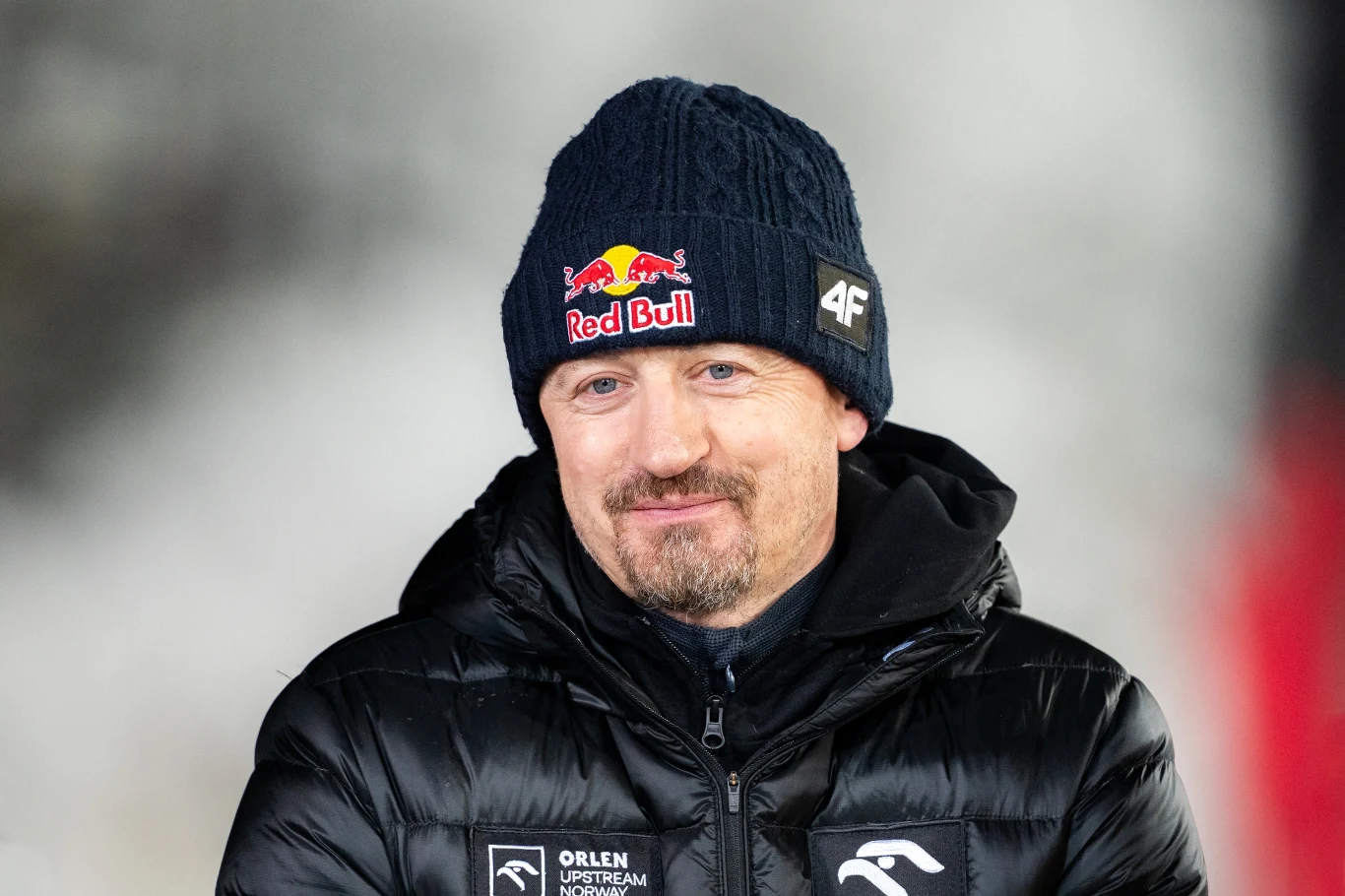mężczyzna w czarnej kurtce zimowej i granatowej czapce z logo Red Bull oraz 4F, delikatnie się uśmiecha, w tle rozmyte jasne i ciemne barwy