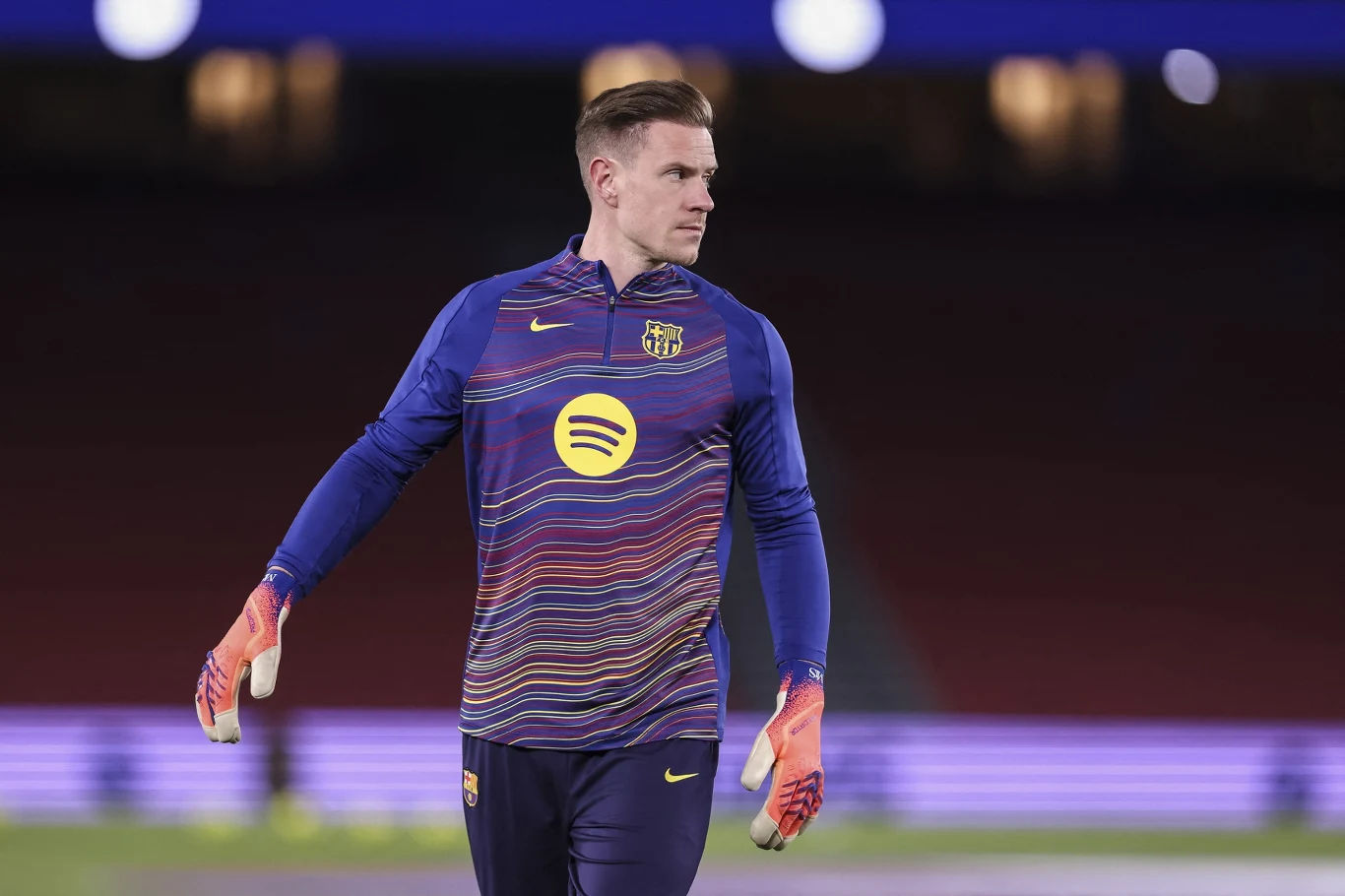 piłkarz w stroju treningowym FC Barcelony ze złotym logotypem Spotify na klatce piersiowej stoi na murawie stadionu, ma na sobie rękawice bramkarskie i patrzy w bok