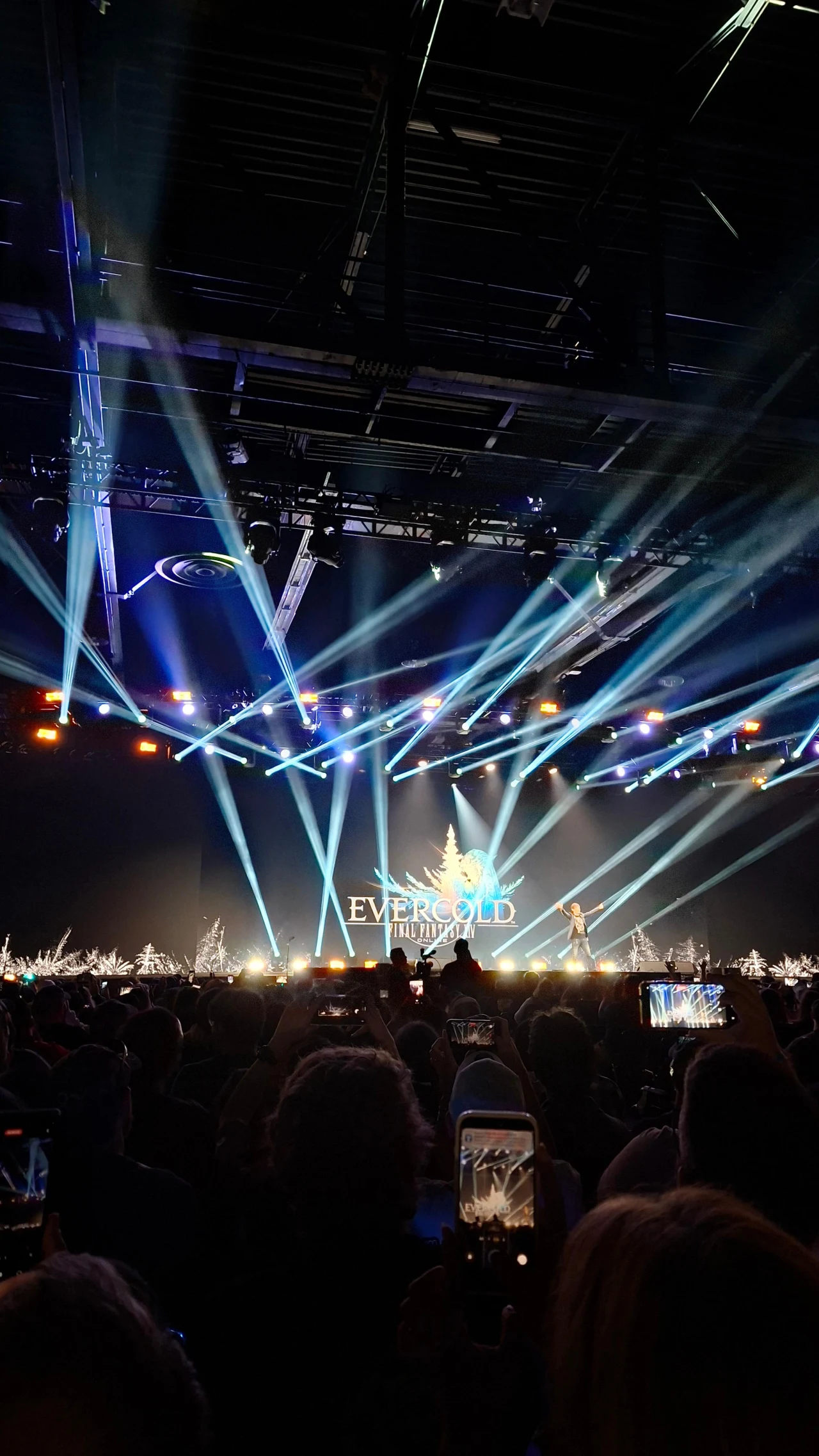źródło: Tomasz Alicki tłum ludzi zgromadzony przed sceną oświetloną intensywnymi reflektorami, na której trwa występ na tle napisu EVERGLOW, widoczne podniesione telefony nagrywające wydarzenie