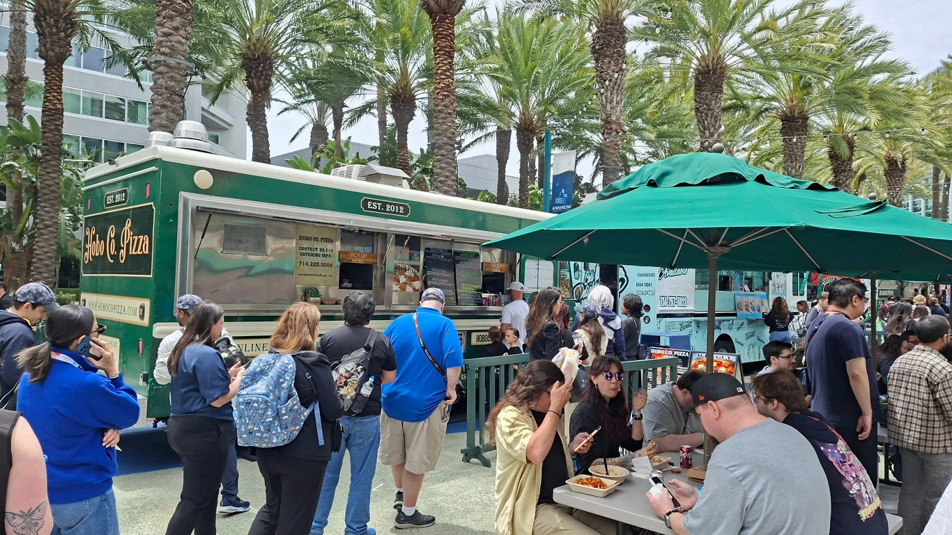 tłum ludzi czekających w kolejce przy food trucku z napojami i pizzą ustawionym w otoczeniu palm, część osób siedzi przy stole pod zielonym parasolem i spożywa jedzenie