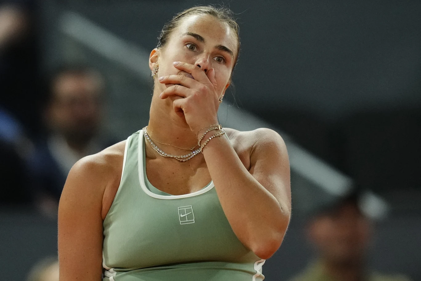 Tenisistka w sportowym stroju z łańcuszkiem na szyi, z ręką przy twarzy w geście zamyślenia lub frustracji, na tle rozmytej publiczności i kortu.