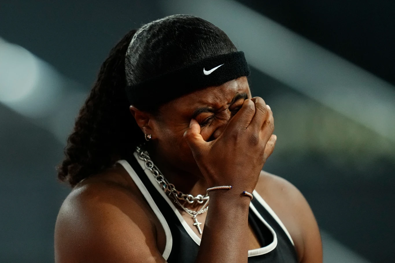 Sportsmenka o ciemnej karnacji z zaciśniętymi oczami, zakrywa twarz dłonią, wyrażając silne emocje; ubrana w czarny top sportowy i opaskę Nike, na szyi srebrny łańcuszek.