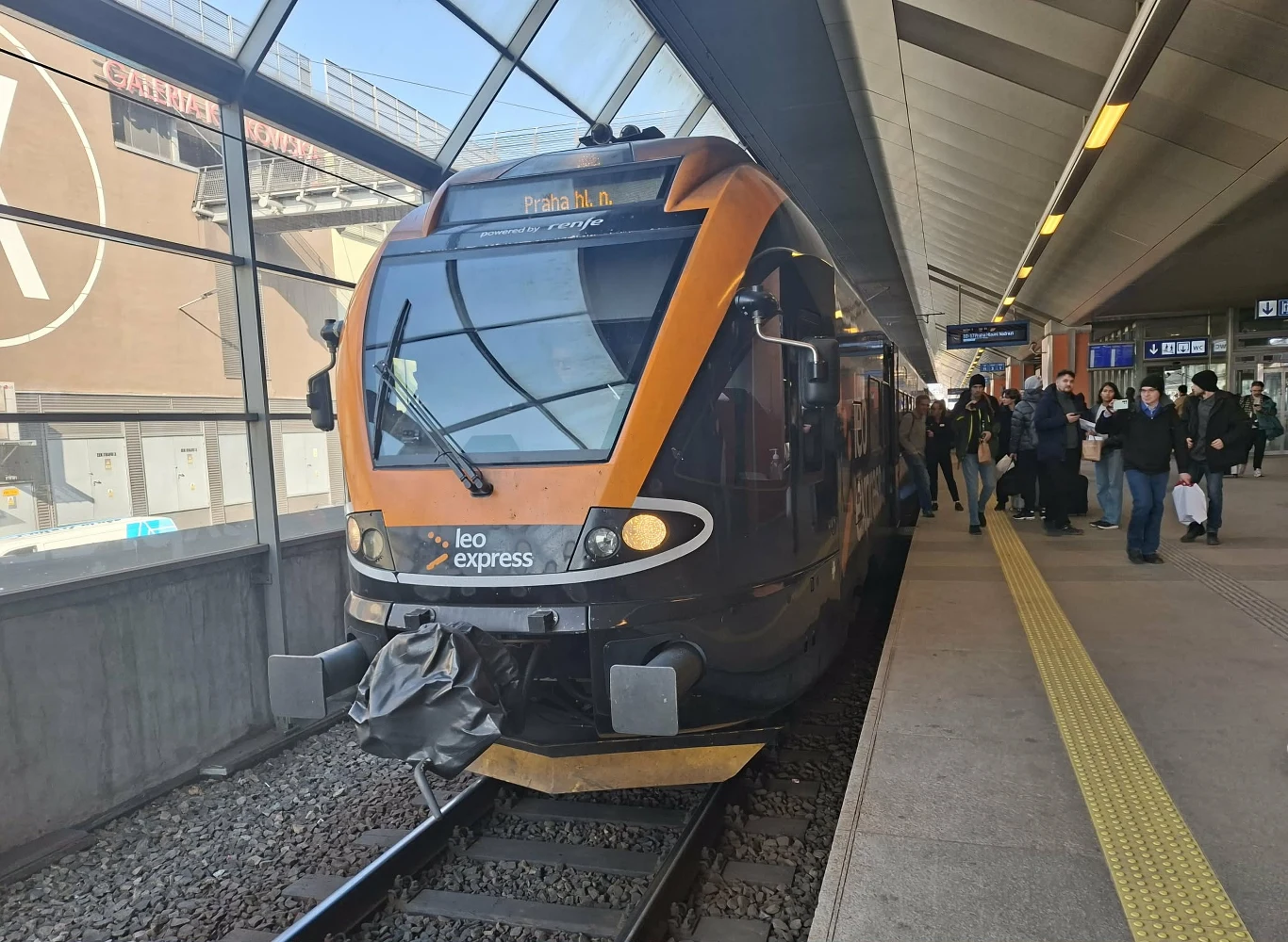 nowoczesny pociąg Leo Express zatrzymany przy peronie, do którego podchodzą pasażerowie z bagażami, jasne i przestronne wnętrze stacji z zadaszeniem oraz oznaczenia informacyjne na peronie