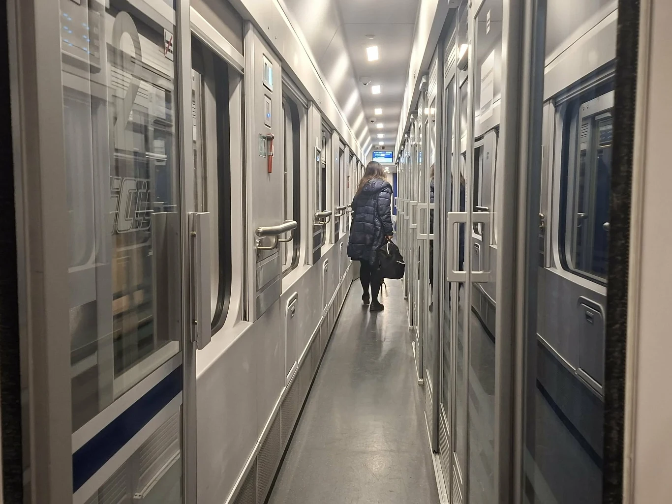 wąski korytarz w wagonie pociągu z osobą w zimowej kurtce idącą w stronę końca wagonu i trzymającą czarną torbę