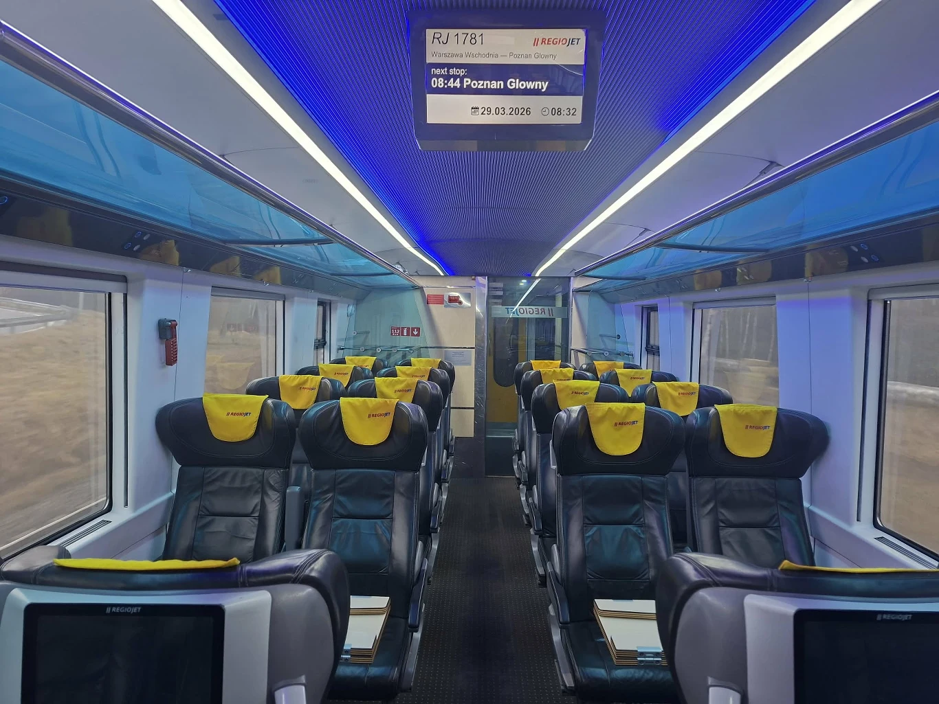 wnętrze nowoczesnego wagonu kolejowego z wygodnymi, rozkładanymi fotelami wyposażonymi w ekrany multimedialne, żółtą tapicerką zagłówków oraz dużymi oknami po bokach; na suficie świetliste niebieskie oświetlenie i elektroniczny wyświetlacz informujący ...