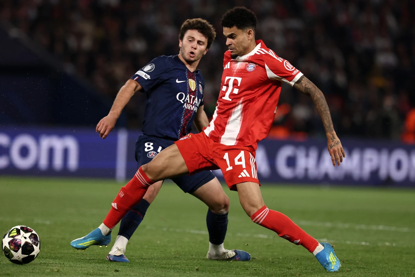 PSG - Bayern Dwóch piłkarzy w trakcie walki o piłkę podczas meczu; zawodnik w czerwono-białym stroju Bayernu Monachium osłania futbolówkę przed rywalem w granatowej koszulce Paris Saint-Germain na zielonym boisku.