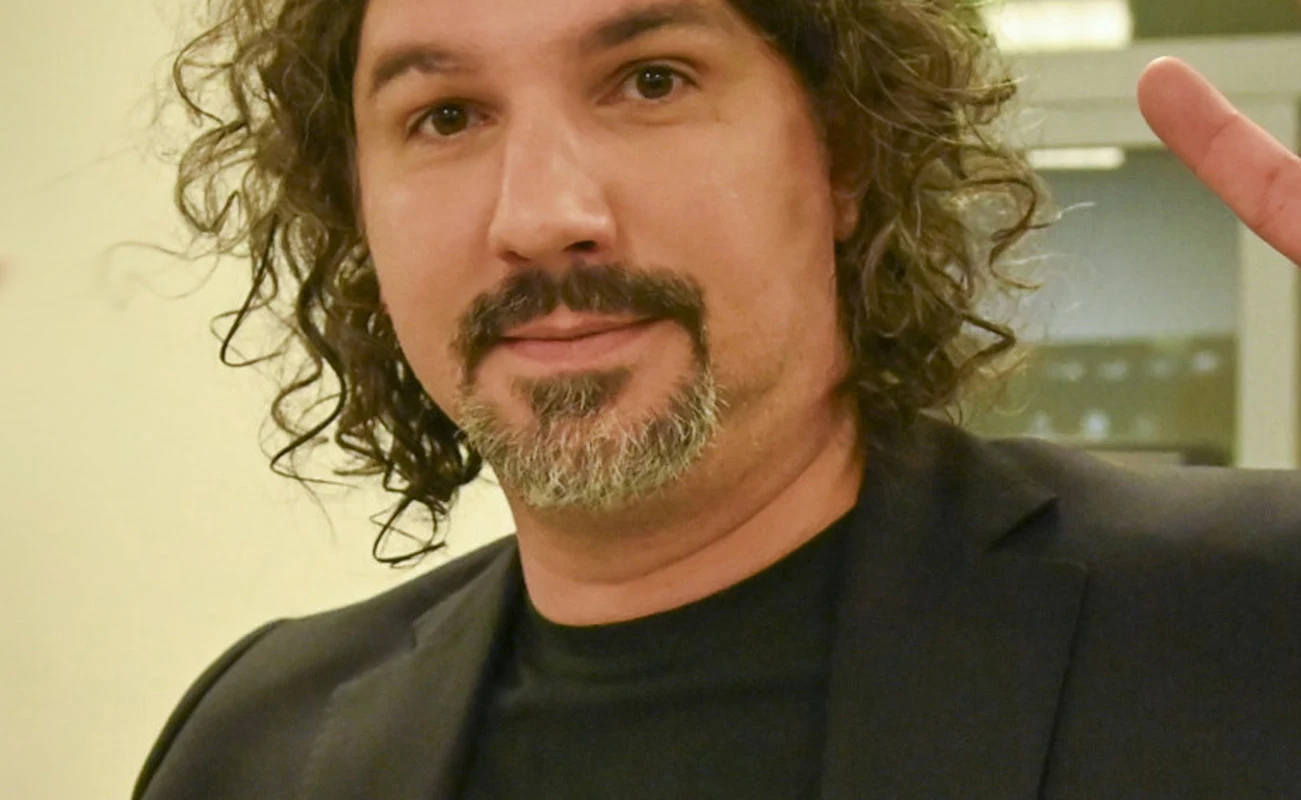 Mężczyzna z kręconymi włosami i zarostem, ubrany w czarną marynarkę i czarną koszulkę, robi znak pokoju dwoma palcami uniesionej ręki.