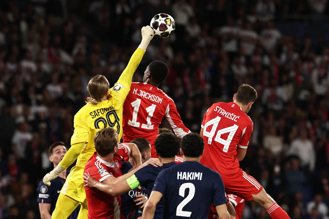 Kadr z wtorkowego starcia gigantów PSG - Bayern bramkarz w żółtym stroju wyskakujący wysoko, wyciąga rękę, aby odbić piłkę podczas walki o futbolówkę z kilkoma obrońcami i napastnikami w czerwonych oraz granatowych koszulkach, pośród tłumu kibiców na trybunach