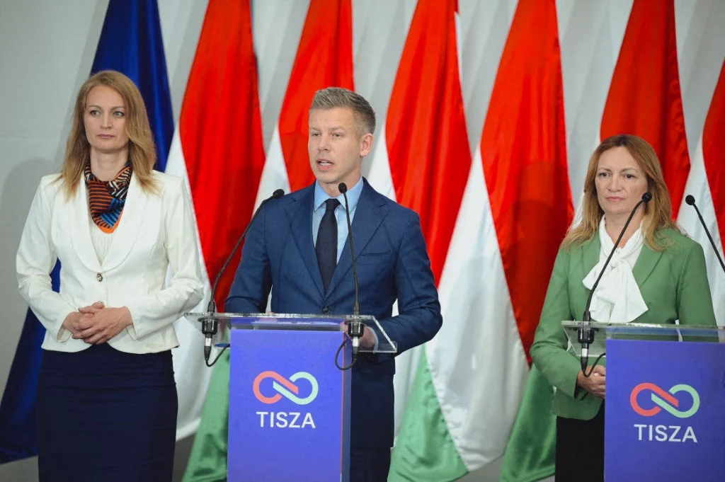 Od lewej: Agnes Forsthoffer, Peter Magyar, Andrea Bujdoso Trzy osoby w eleganckich strojach stoją za mównicami na tle biało-czerwono-zielonych flag oraz symbolu i napisu TISZA, centralna postać przemawia do mikrofonu.