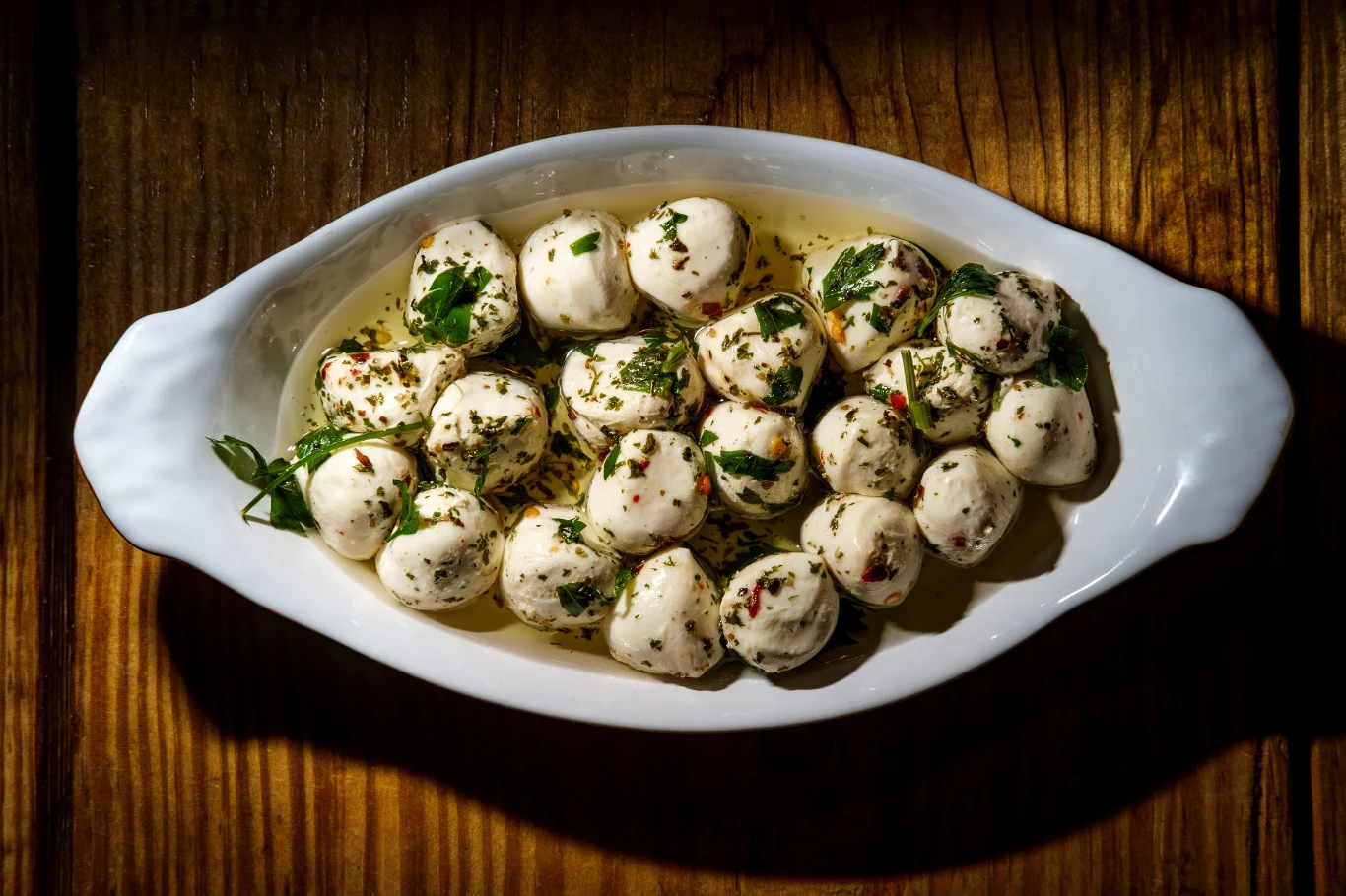 Mozzarella z aromatycznymi dodatkami świetnie uzupełni grillowe przysmaki Kuleczki mozzarelli zanurzone w oliwie z oliwek, posypane świeżymi ziołami, płatkami chili oraz listkami pietruszki, podane w białej, owalnej misce na drewnianym stole.