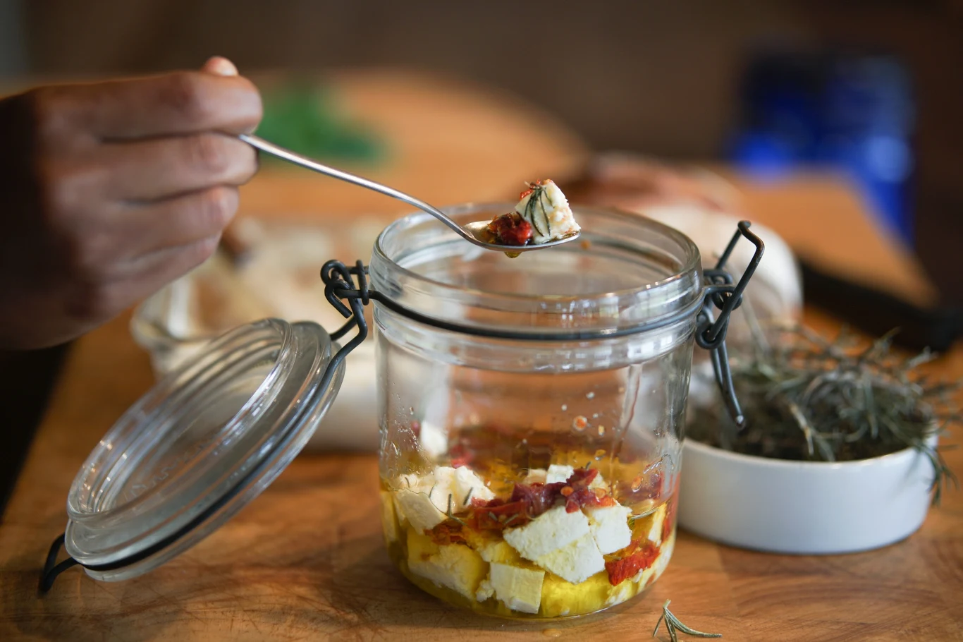 Mozzarella, ogórki i pomidorki w zupełnie nowej odsłonie. Zamknij w słoikach i zabierz na grilla, a oczarujesz wszystkich smakoszy Słoik z marynowaną fetą, suszonymi pomidorami i oliwą, obok miseczka ziołami i ręka z łyżką.