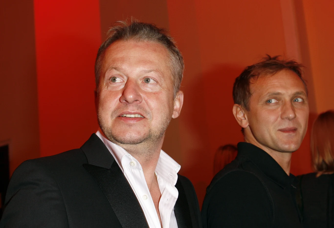 Bogusław Linda i Andrzej Chyra Bogusław Linda i Andrzej Chyra w eleganckich marynarkach, stojący obok siebie w czerwonym oświetleniu.
