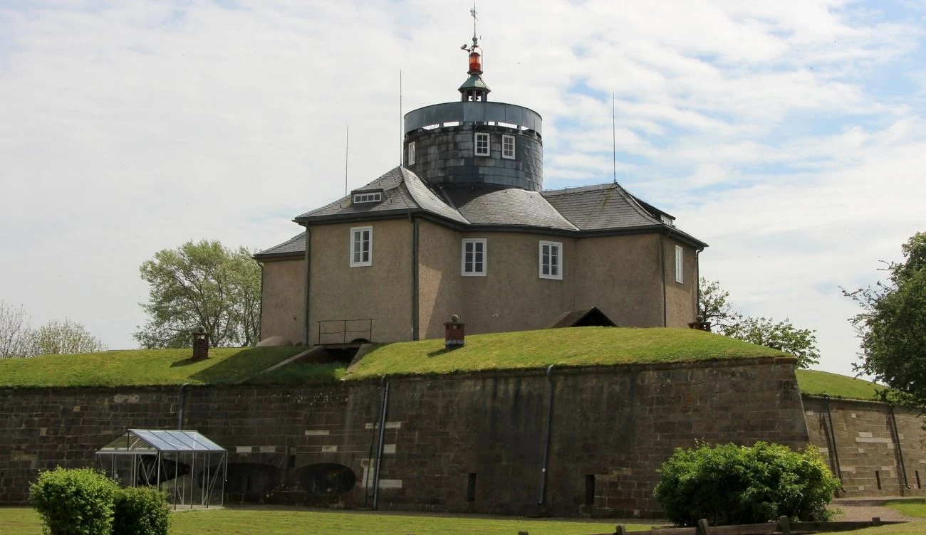 okrągły budynek obserwatorium astronomicznego z jasną elewacją i licznymi oknami, umieszczony na szczycie solidnego, kamiennego bastionu otoczonego zielonym trawnikiem, z kopułą i wieżyczką na dachu oraz metalową konstrukcją po lewej stronie w tle