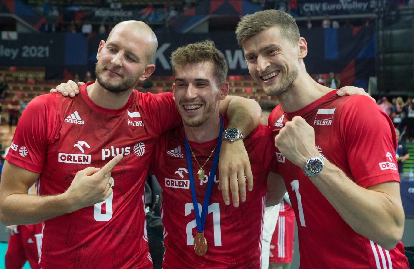Piotr Nowakowski (pierwszy z prawej) w reprezentacji Polski trzech siatkarzy w czerwonych koszulkach reprezentacji Polski stoi obok siebie, uśmiecha się i obejmuje, środkowy zawodnik ma na szyi brązowy medal, wszyscy wyglądają na zadowolonych i dumni z osiągnięcia