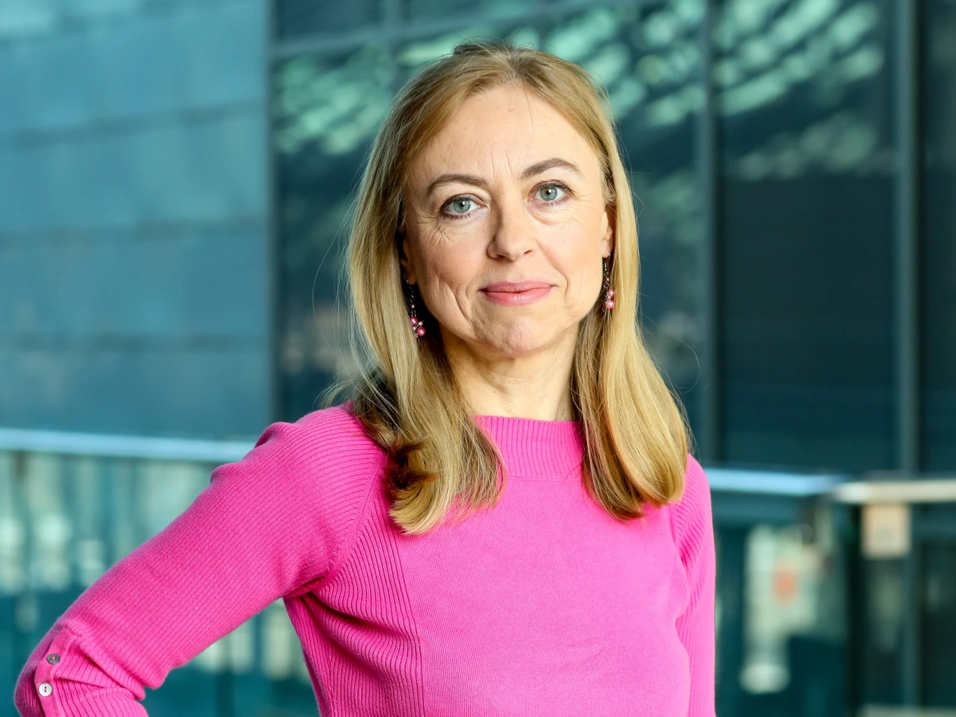 Monika Rosca w 2015 roku Kobieta o blond włosach ubrana w różowy sweter, stojąca w nowoczesnym wnętrzu, patrząca z lekkim uśmiechem