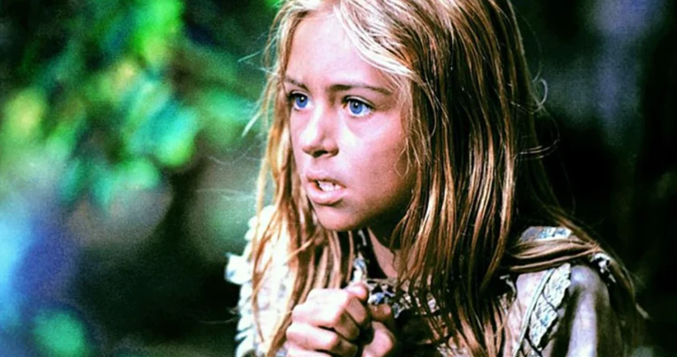 Kadr z filmu "W pustyni i w puszczy" (1973) Dziewczynka o jasnych włosach i niebieskich oczach, z wyrazem determinacji na twarzy oraz splecionymi przed sobą dłońmi, stojąca na tle rozmytego, zielonego otoczenia.