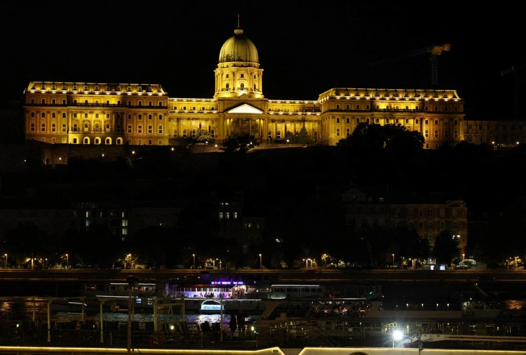 Wzgórze Zamkowe w Budapeszcie oświetlony pałac z dużą kopułą centralną dominuje nad ciemnym otoczeniem, poniżej widoczna jest rzeka z przystanią i zacumowanymi statkami