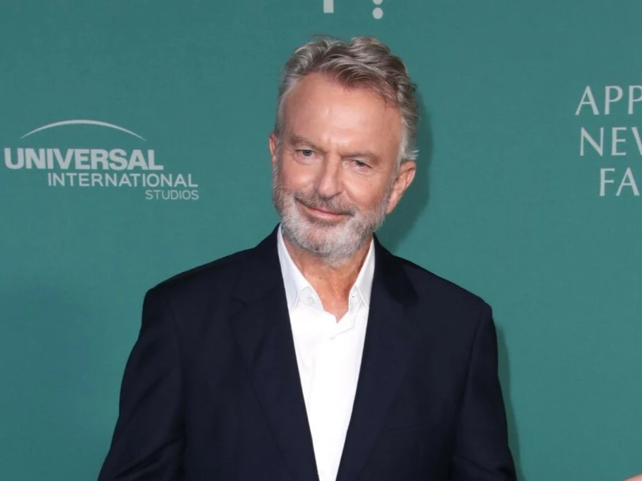 Sam Neill Mężczyzna z siwą brodą i krótkimi włosami ubrany w ciemny garnitur oraz białą koszulę stoi na tle zielonej ścianki.