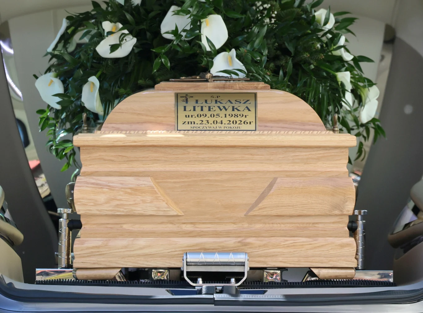 drewniana trumna z tabliczką imienną oraz datami narodzin i śmierci, ustawiona w samochodzie pogrzebowym, ozdobiona bukietem białych kwiatów i zielonych liści