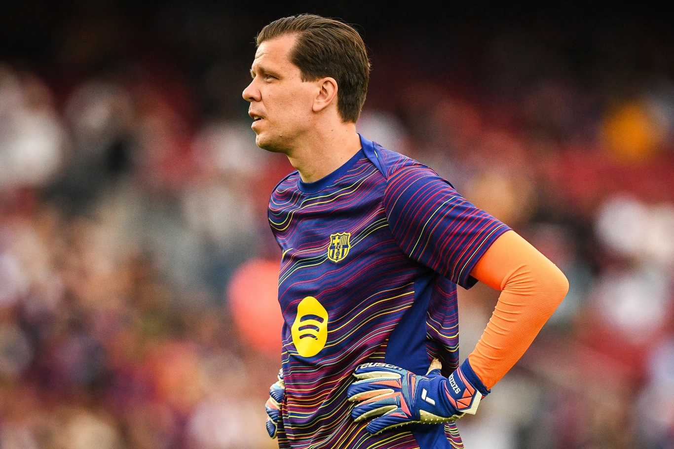 Wojciech Szczęsny Wojciech Szczęsny w pomarańczowej koszulce bramkarskiej stoi na tle trybun stadionu.
