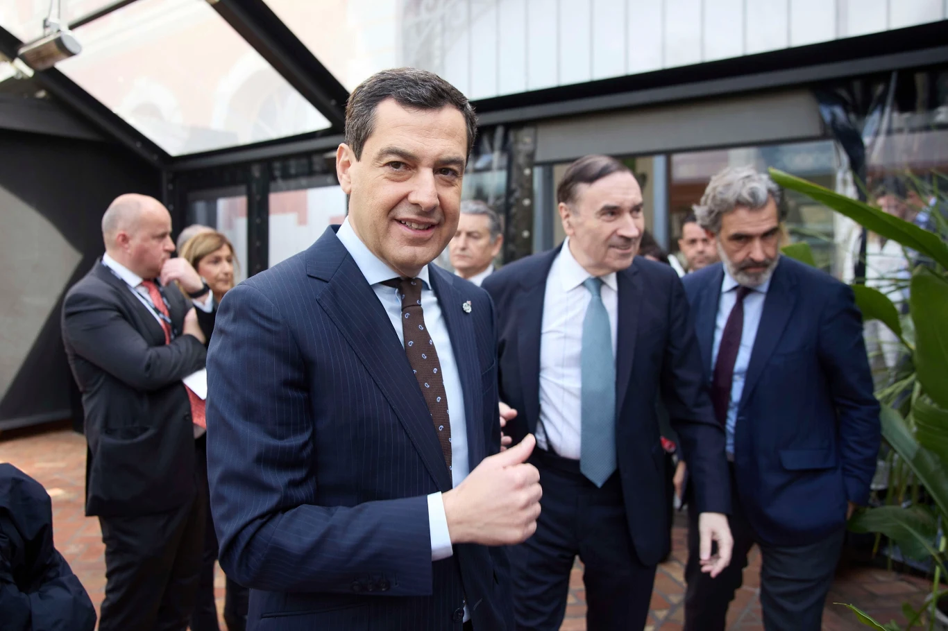 Mężczyzna w garniturze z uśmiechem i uniesionym kciukiem stoi na pierwszym planie, za nim grupa elegancko ubranych osób rozmawiających w przeszklonym pomieszczeniu.