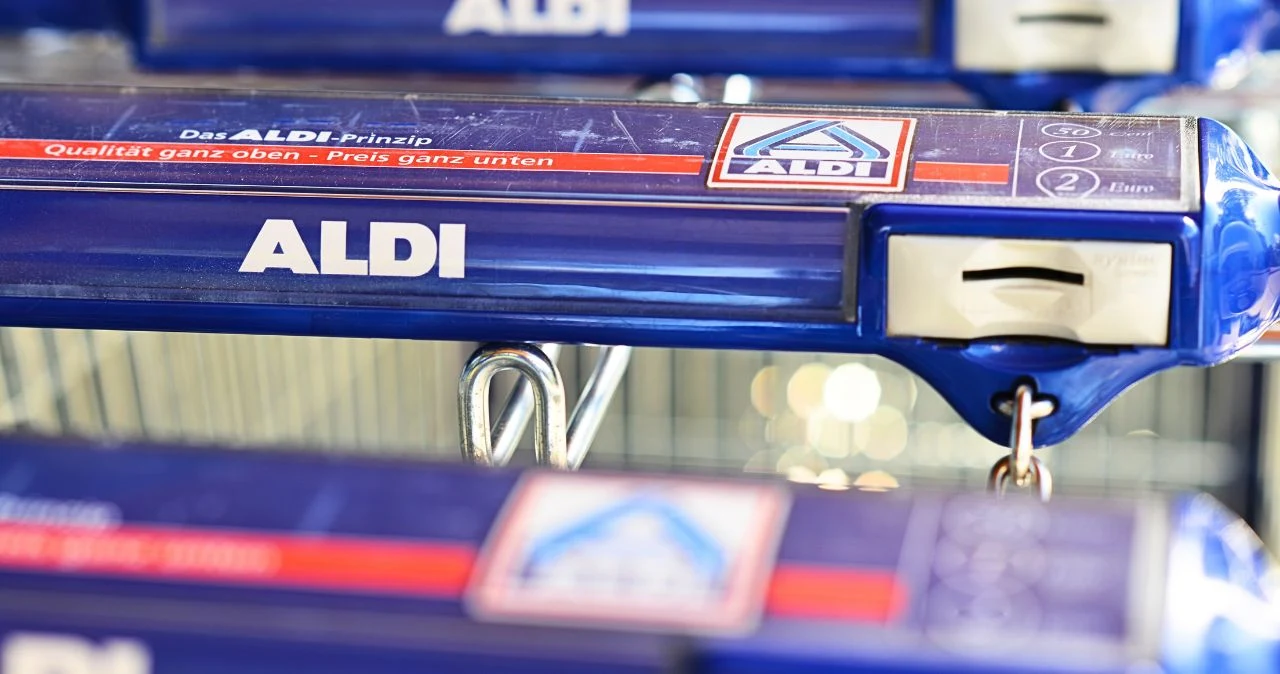 ALDI odpaliło mocne promocje – nawet do 90% taniej. Sprawdź, co warto kupić z nowej gazetki Zbliżenie na niebieski uchwyt wózka sklepowego ALDI z białym logo i mechanizmem na monetę, w tle inne wózki.