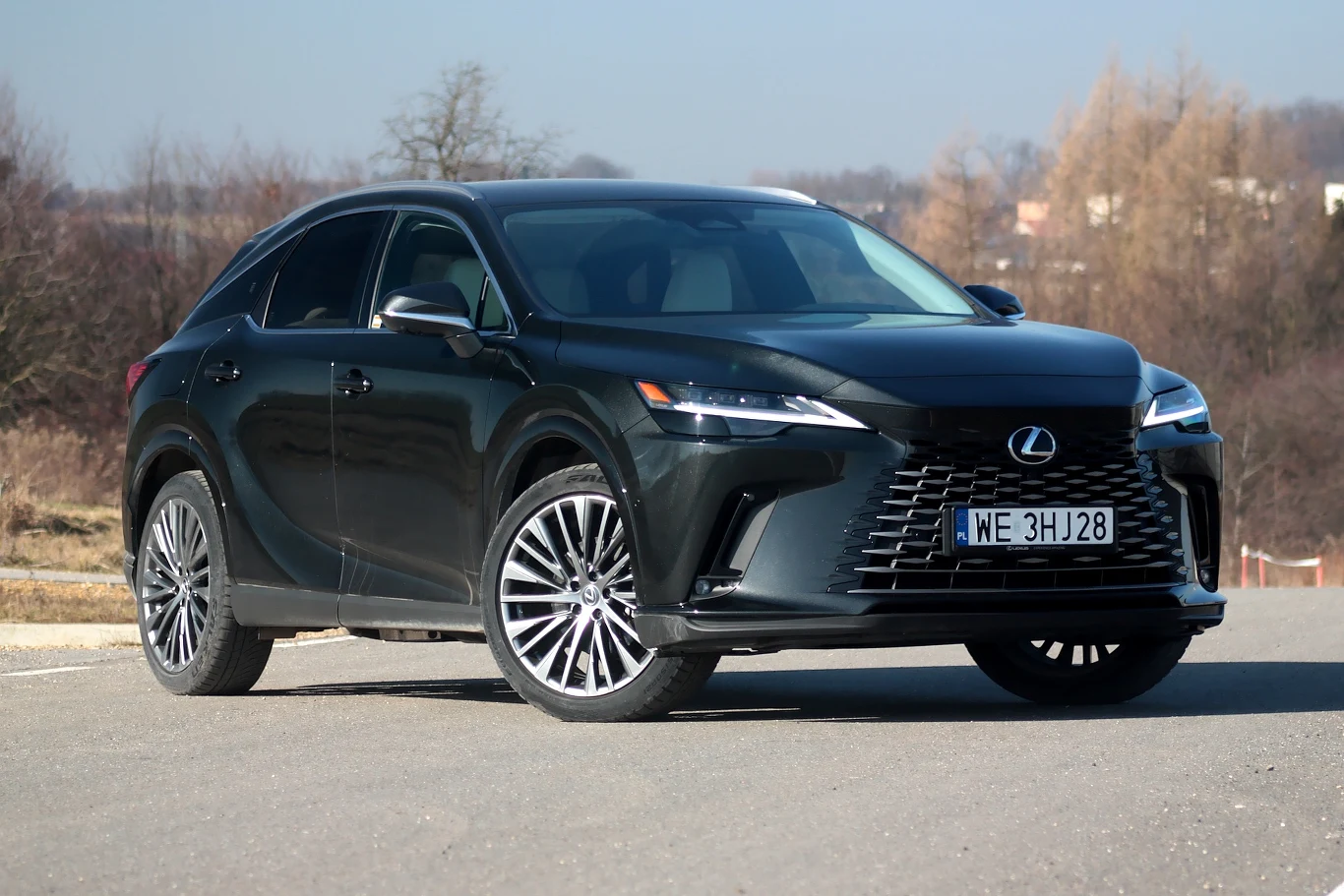 czarny SUV marki Lexus z wyrazistym grillem i chromowanymi detalami, ustawiony na asfaltowej nawierzchni w otoczeniu drzew bez liści i jasnego nieba